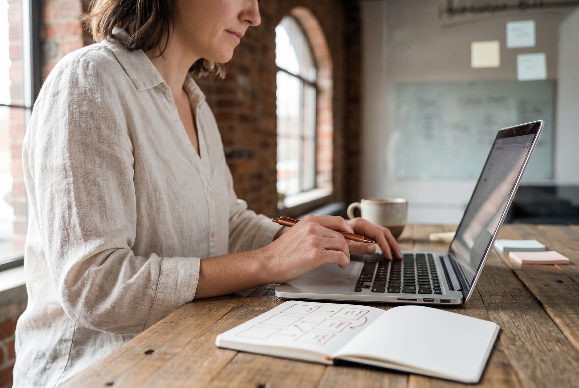 Close-up of a workshop participant in a linen shirt working at a laptop, holding a copper-coloured fineliner over an open notebook that shows simple boxes and arrows sketched in copper ink — a Tabakfabrik-style industrial workspace in the soft background.